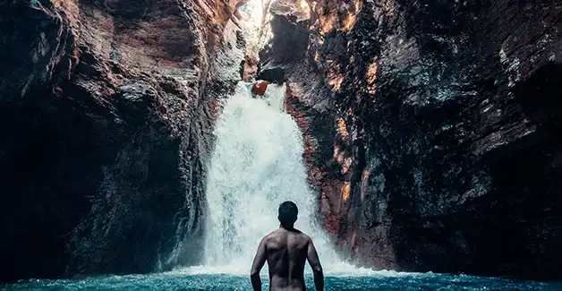 A man standing in the water in front of a waterfall.